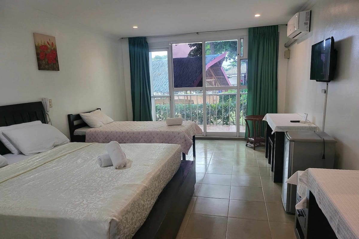 Habitación de hotel con dos camas, un escritorio, televisor y ventana con vistas a edificios y vegetación.