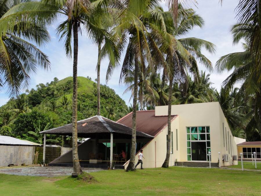 A building with a sloped roof and a covered outdoor area, surrounded by palm trees and greenery.