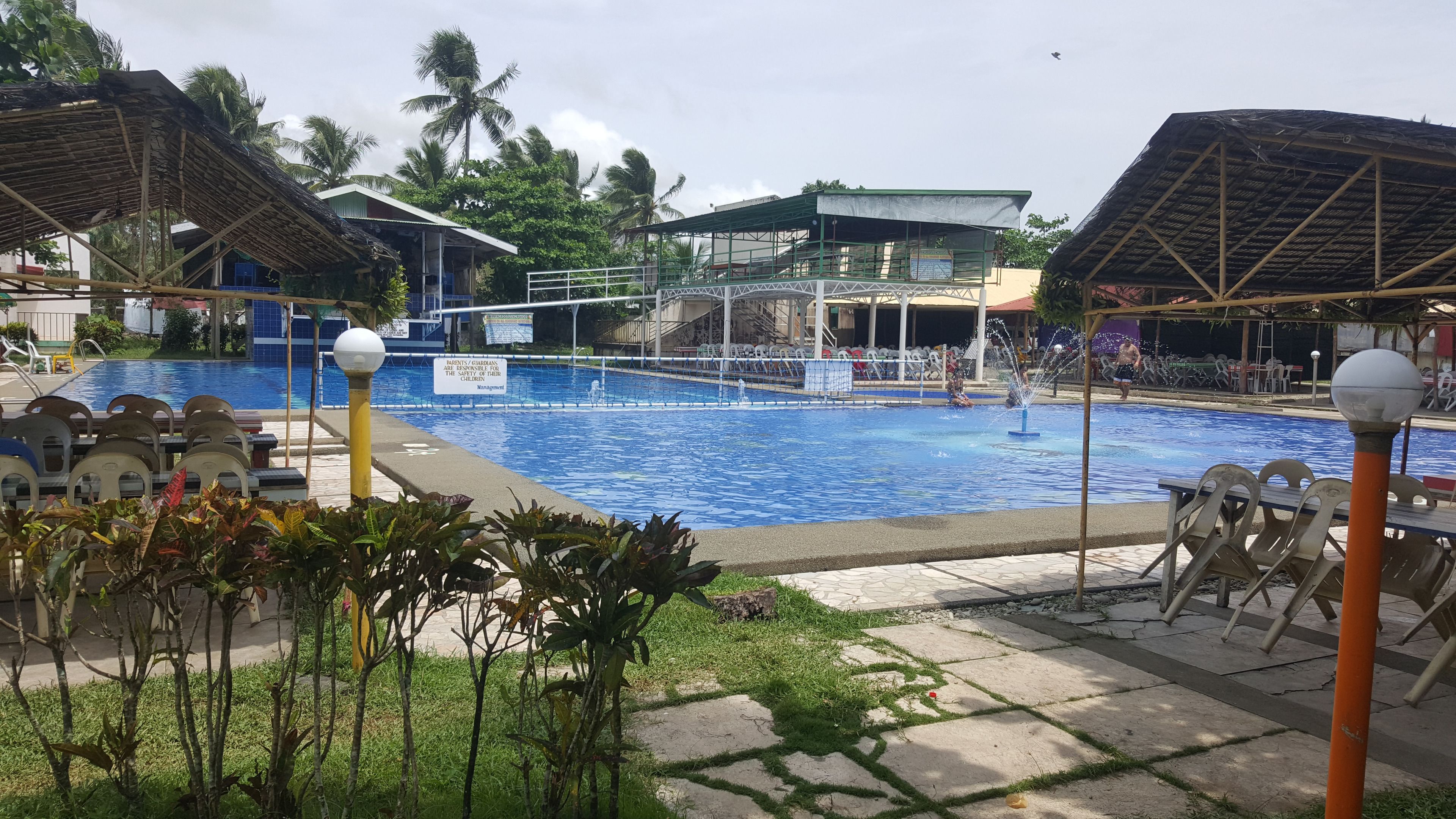 A swimming pool with a sign indicating responsibility for children, surrounded by palm trees and covered structures.