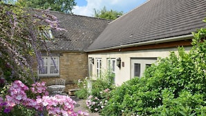 Ein Haus aus Stein mit einem Ziegeldach, ein kleiner Garten mit rosa Blumen und ein Metallstuhl.