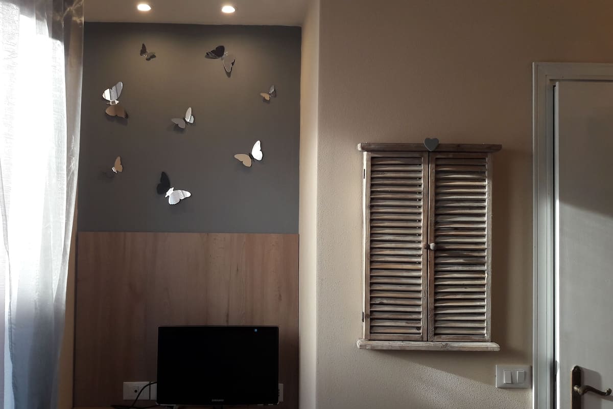 A room with a wall-mounted TV, wooden paneling, a window with shutters, and butterfly wall art.