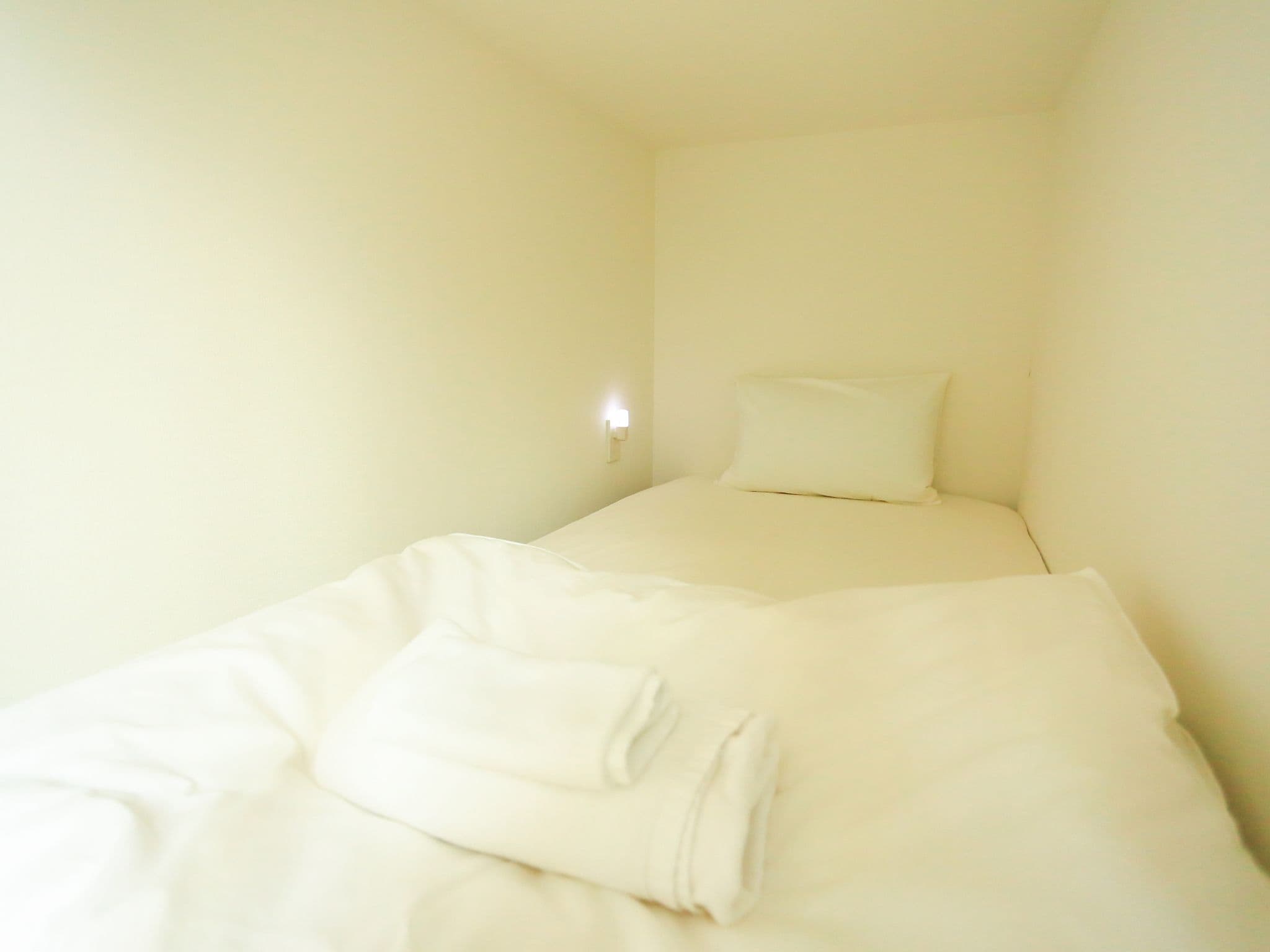 A small, single-bed room with a plain white wall and a bedside lamp.