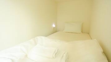 A small, single-bed room with a plain white wall and a bedside lamp.