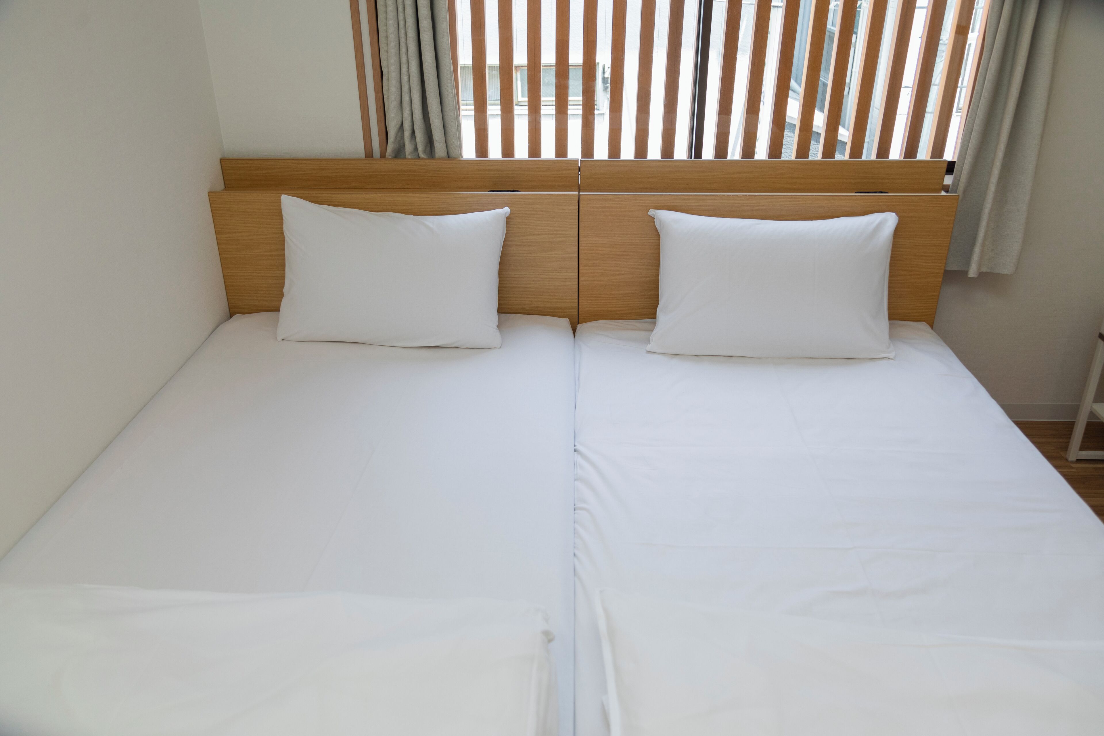A double bed with a wooden headboard, white bedding, and a view of the city through vertical blinds.