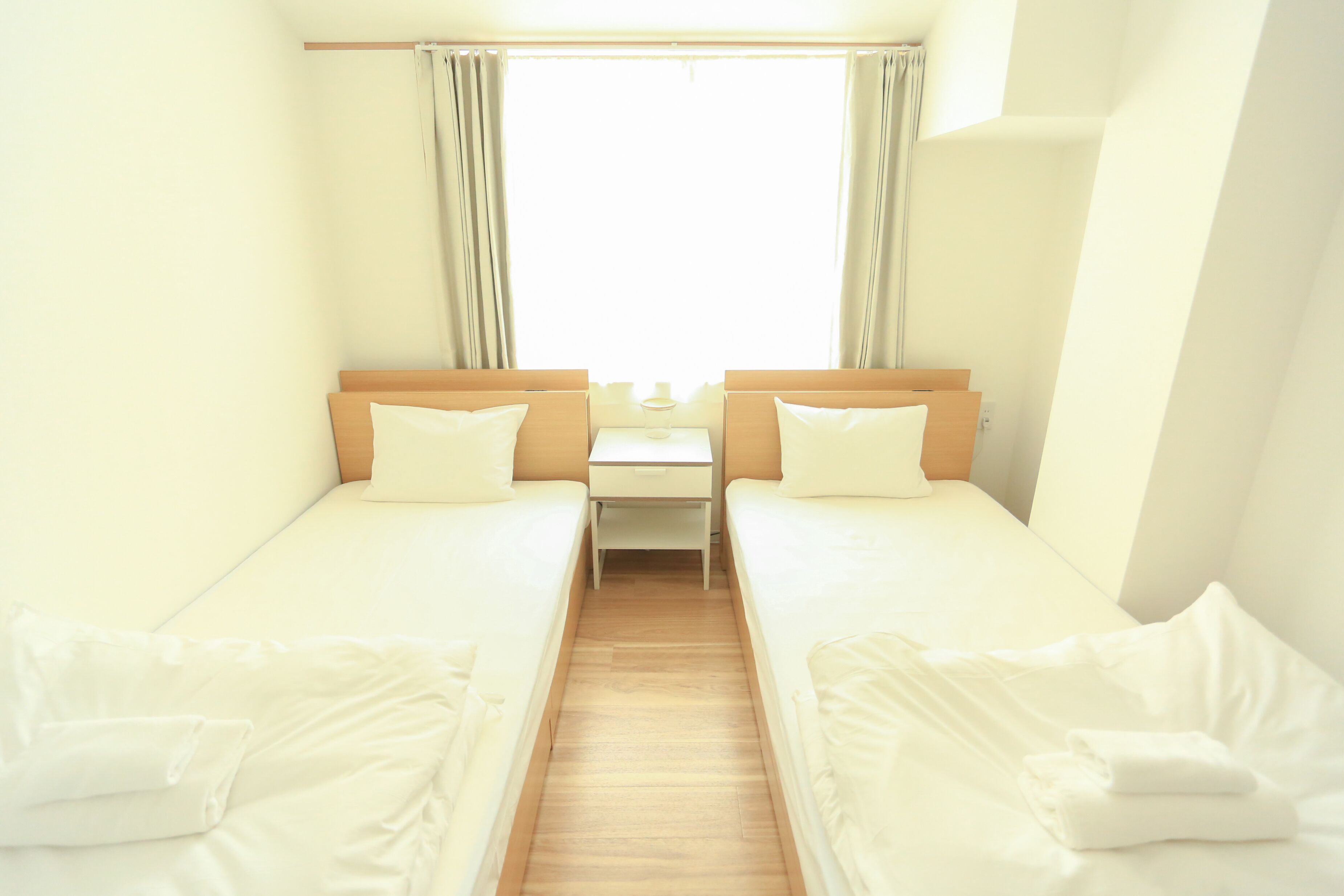 Una habitación pequeña y limpia con dos camas individuales, una mesita y una ventana con cortinas.
