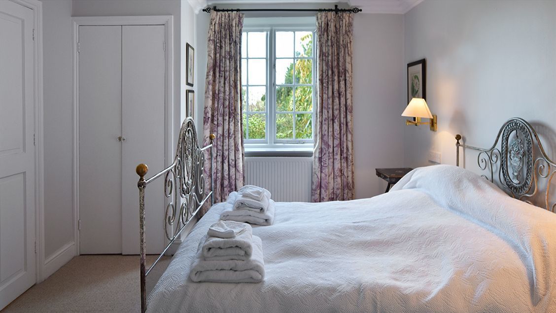 A neatly made bed with folded towels, a window with curtains, a bedside lamp, and a framed picture on the wall.