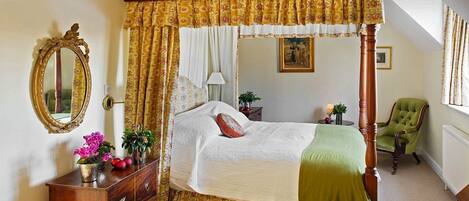 A four-poster canopy bed with a wooden headboard, a green armchair, a wooden dresser with decorative items, and a mirror on the wall.