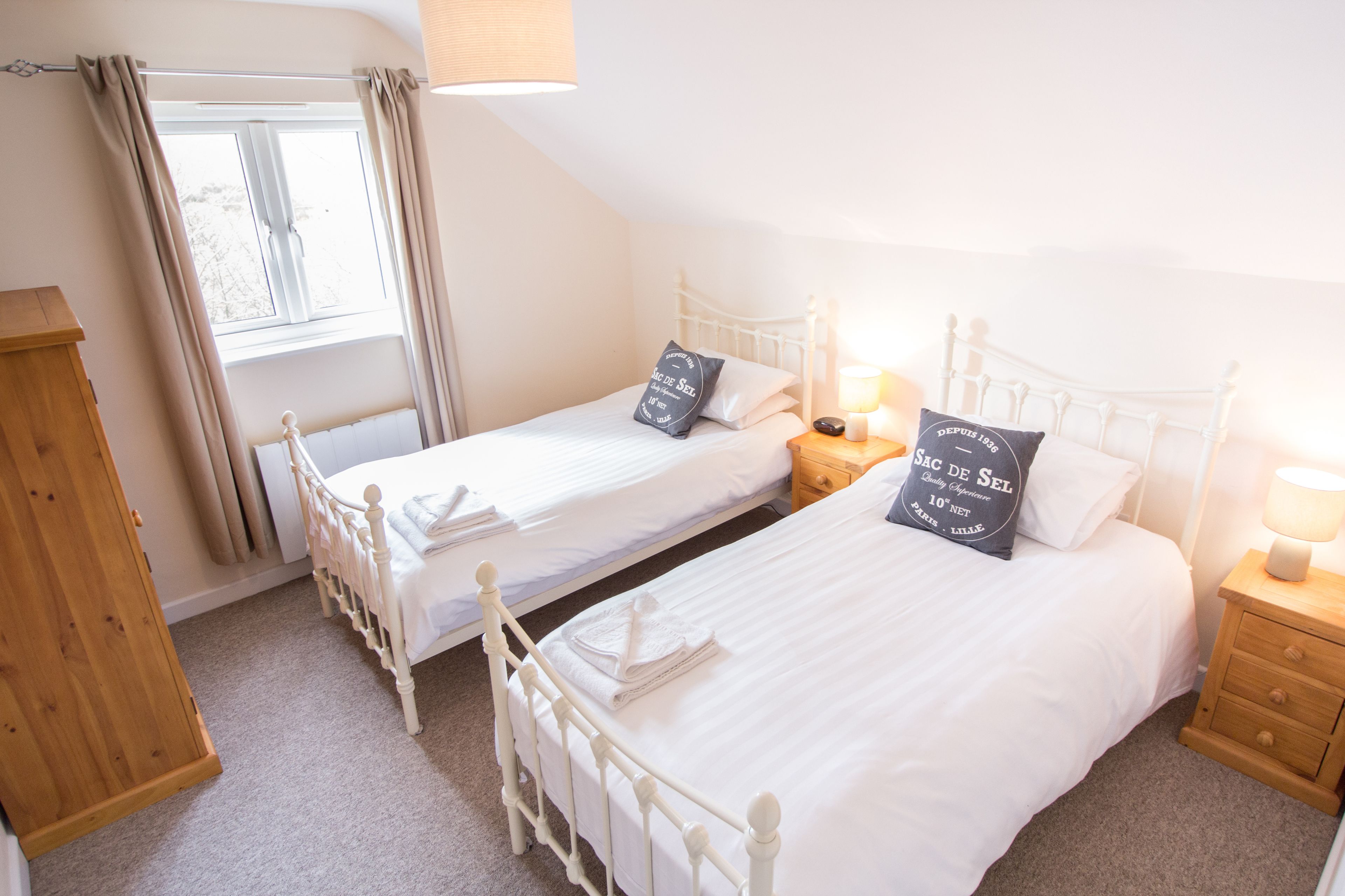 A bedroom with two single beds, a wooden wardrobe, a window with curtains, and bedside lamps.