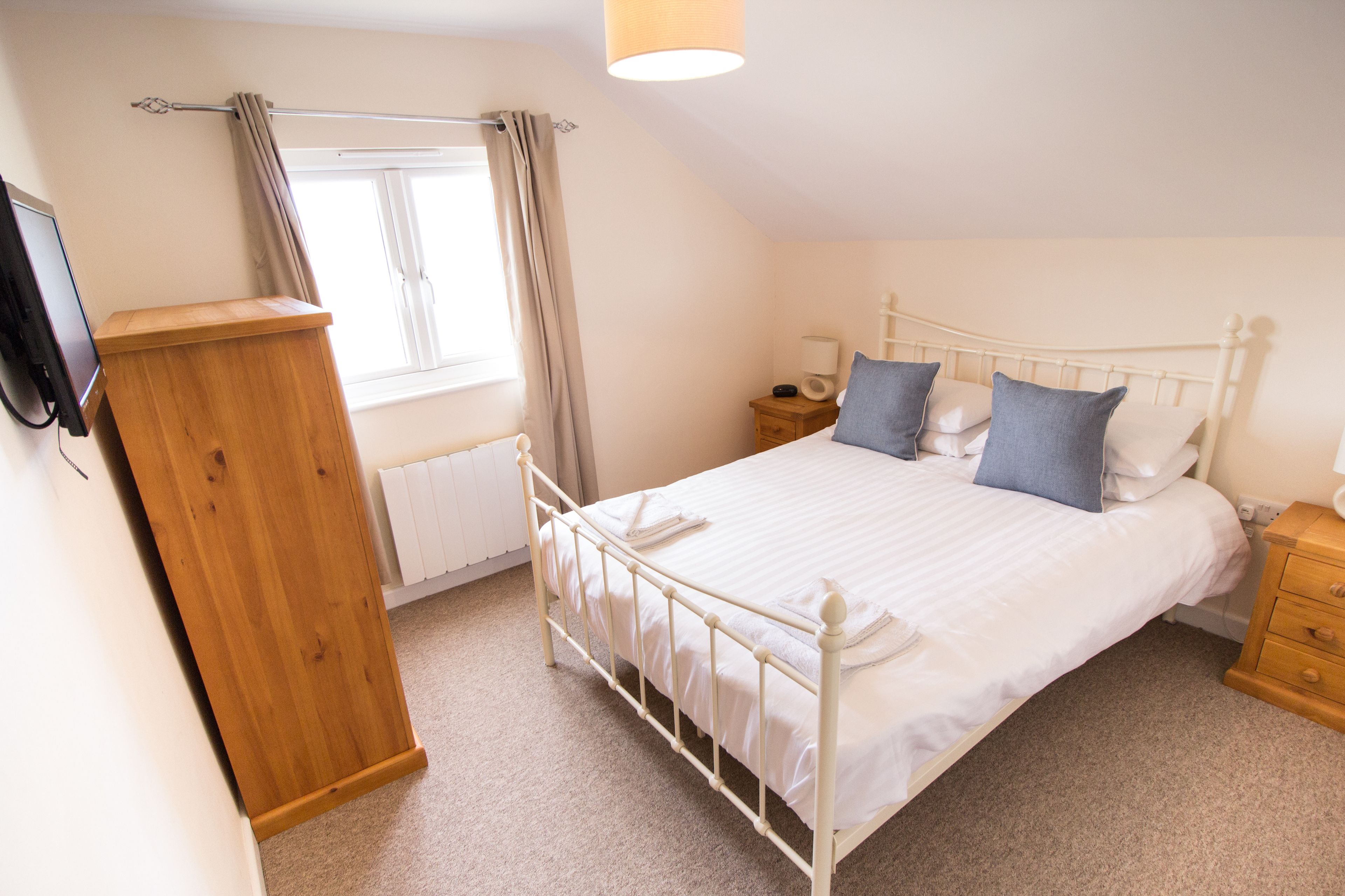 A bedroom with a bed, wooden dresser, bedside table, lamp, and a window with curtains.