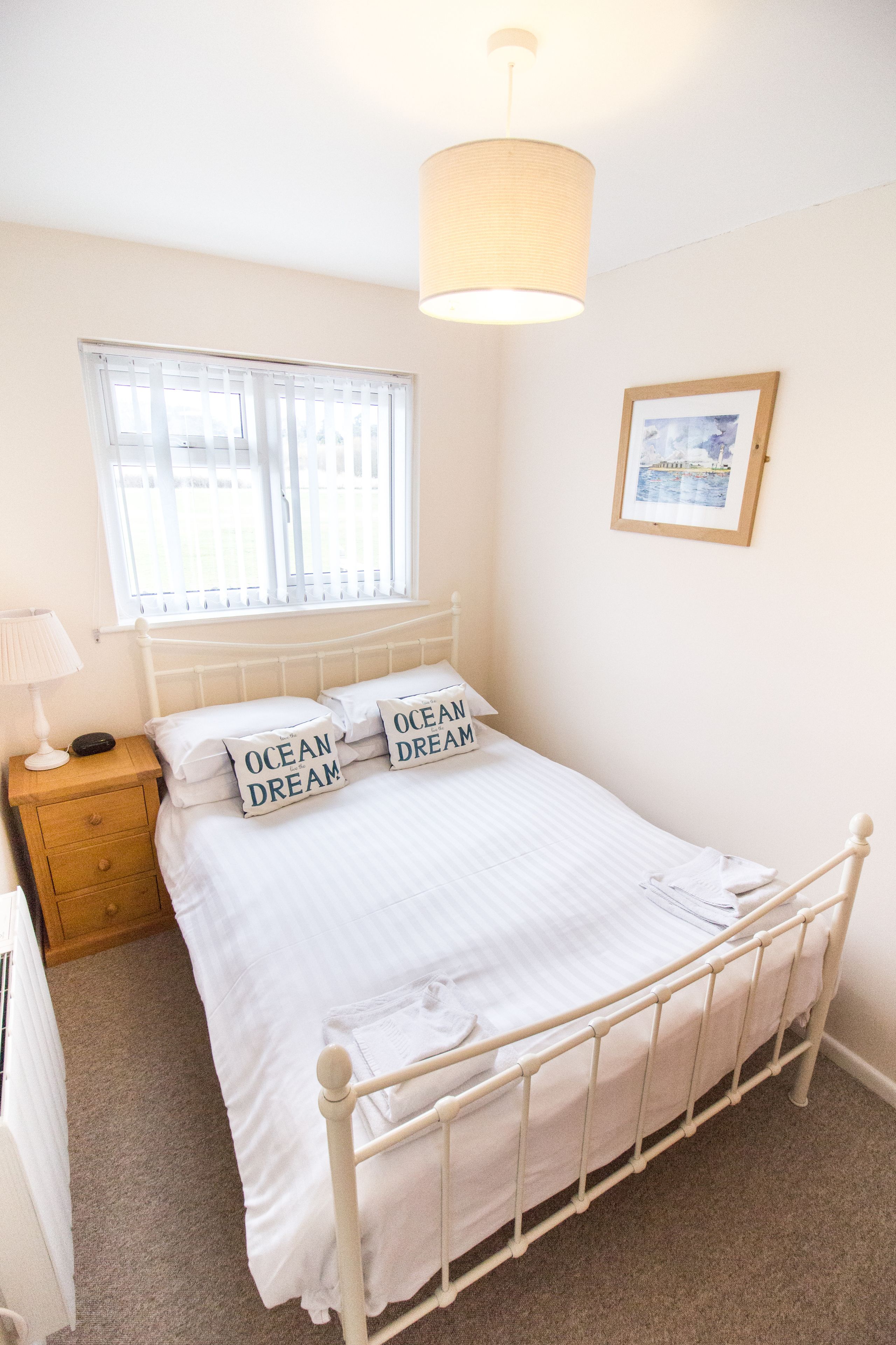 A neatly made bed with white bedding and pillows, a bedside table with a lamp, a window with blinds, and a framed picture on the wall.