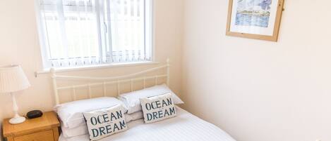 Un lit bien fait, avec du linge de lit et des oreillers blancs, une table de chevet avec une lampe, une fenêtre avec des stores et un tableau encadré au mur.