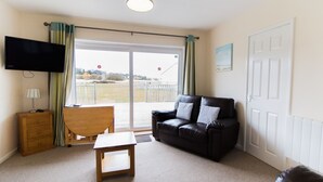 A living room with a black leather sofa, a wooden table, a TV mounted on the wall, and a sliding glass door leading to an outdoor area. - Seaview Holidays - Salterns Village (Seaview)