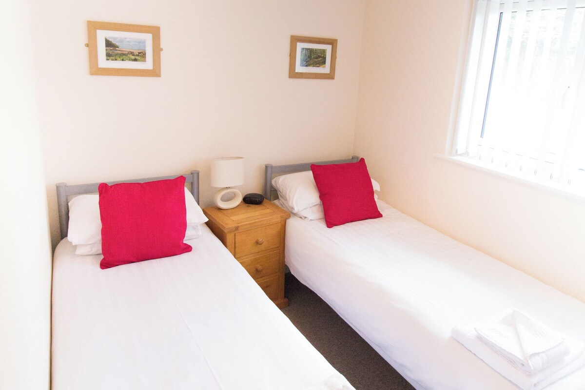 Une petite chambre propre, avec deux lits simples, une table de chevet avec une lampe et une fenêtre équipée de stores.