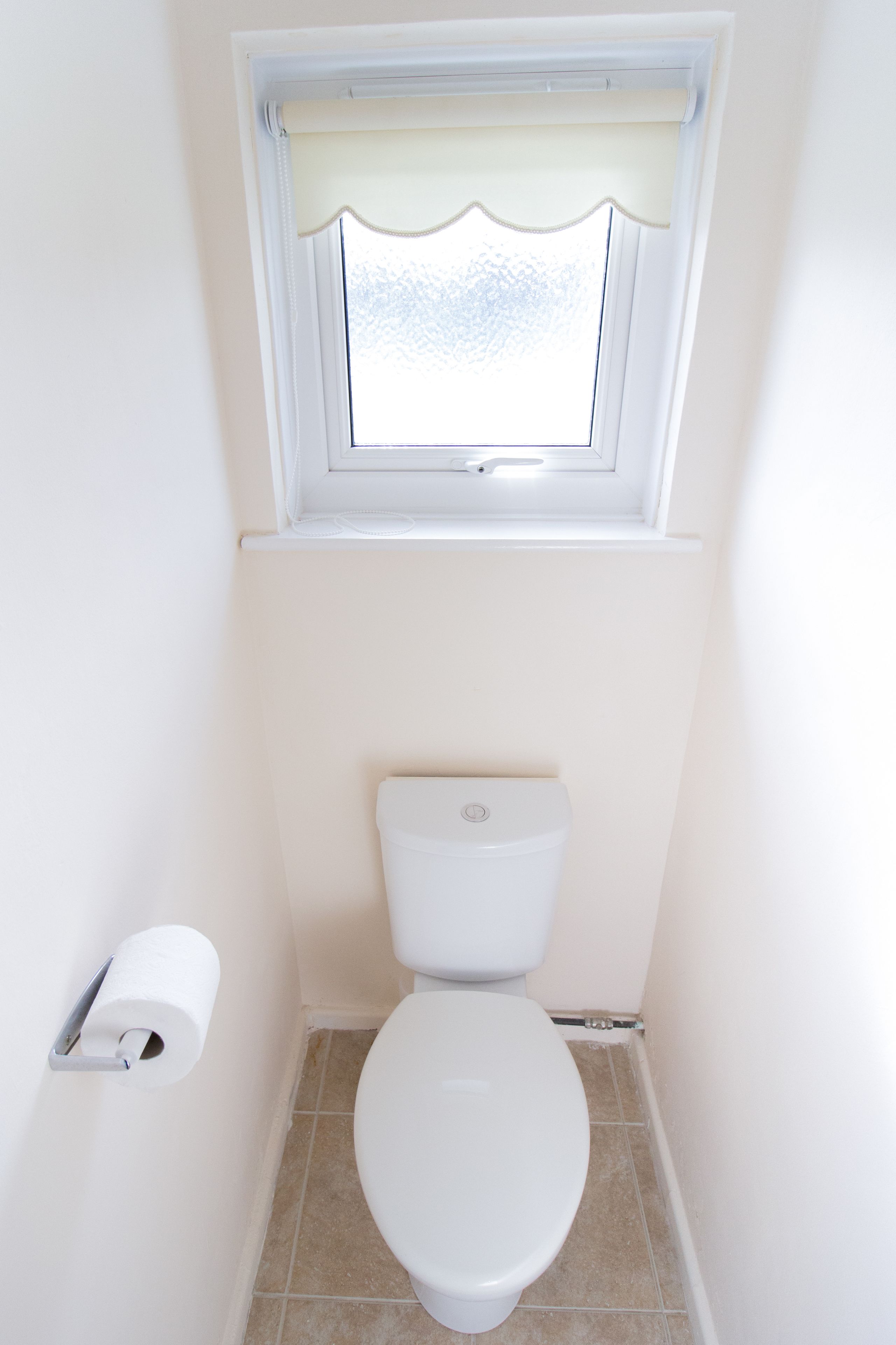 Ein kleines, weißes Badezimmer mit Toilette, einem Fenster mit weißem Vorhang und einer Toilettenpapierrolle an der Wand.