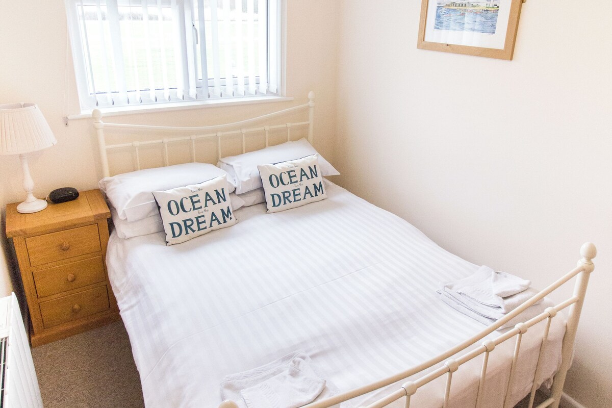 A neatly made bed with white bedding and pillows, a bedside table with a lamp, a window with blinds, and a framed picture on the wall.