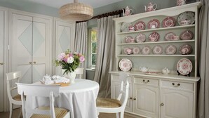 A dining area with a round table, white chairs, a vase with flowers, and a china cabinet with plates and teapots. - Cheltenham Cottage (Chipping Norton)