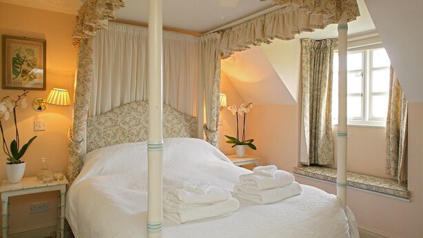 A four-poster bed with a canopy, a bedside table with a lamp, a potted plant, and a window with curtains. - Cheltenham Cottage (Chipping Norton)