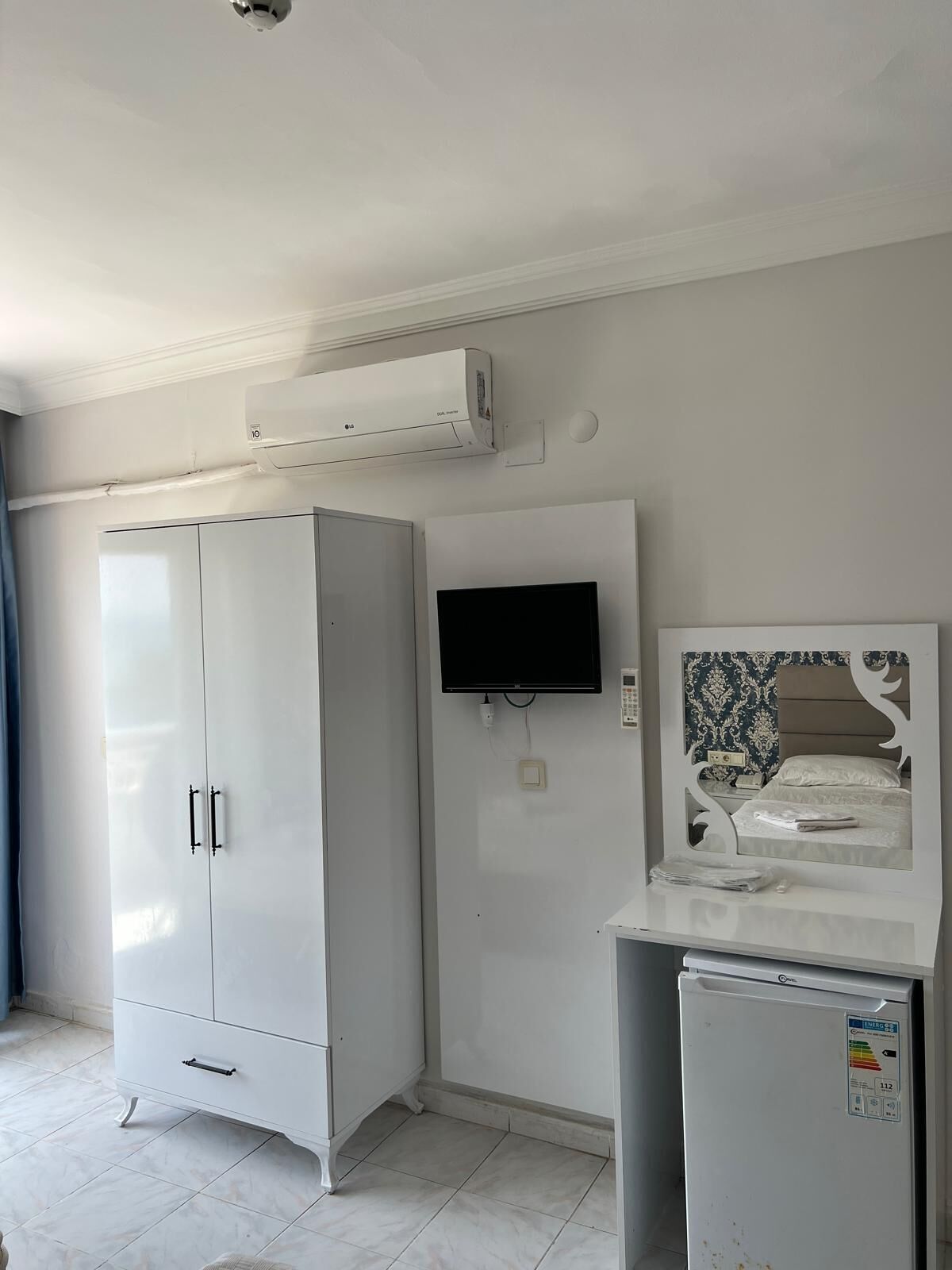 Una habitación con un armario blanco, un televisor de pantalla plana montado en la pared y un refrigerador pequeño.