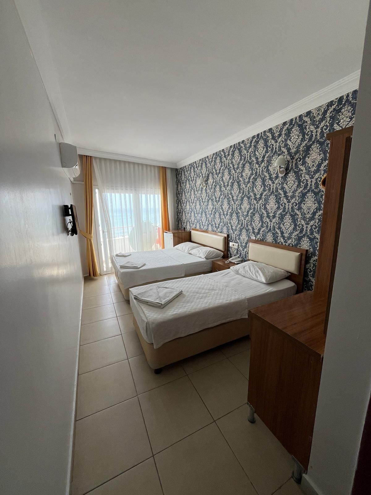 A hotel room with two beds, a wooden desk, a patterned wallpaper, and a window with curtains.
