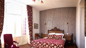 A bedroom with a large bed, a red chair, a curved wall, and a window with curtains. - Villa L'Orante (Lourdes)