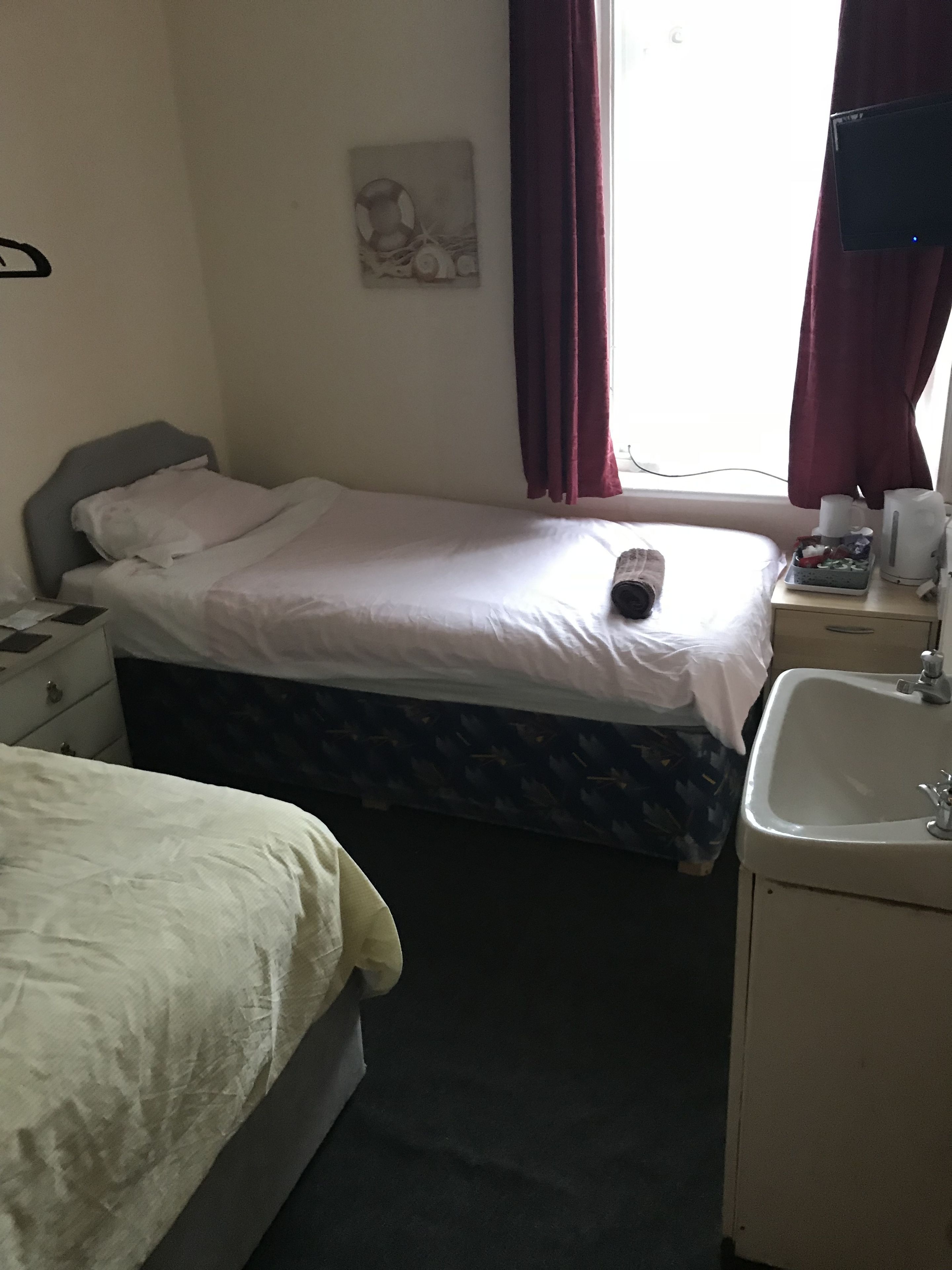 A small hotel room with two single beds, a bedside table with a lamp, a small sink, and a window with curtains.