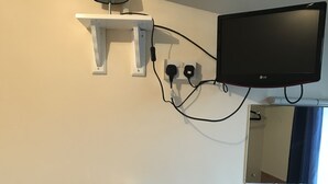 Casa de banho com lavatório branco, espelho, prateleira com chaleira e chávena, e televisão na parede.