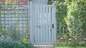 A paved pathway leading to a white gate with vertical bars, surrounded by lush greenery and a brick building in the background. - Hedingham Old Pottery (Halstead)