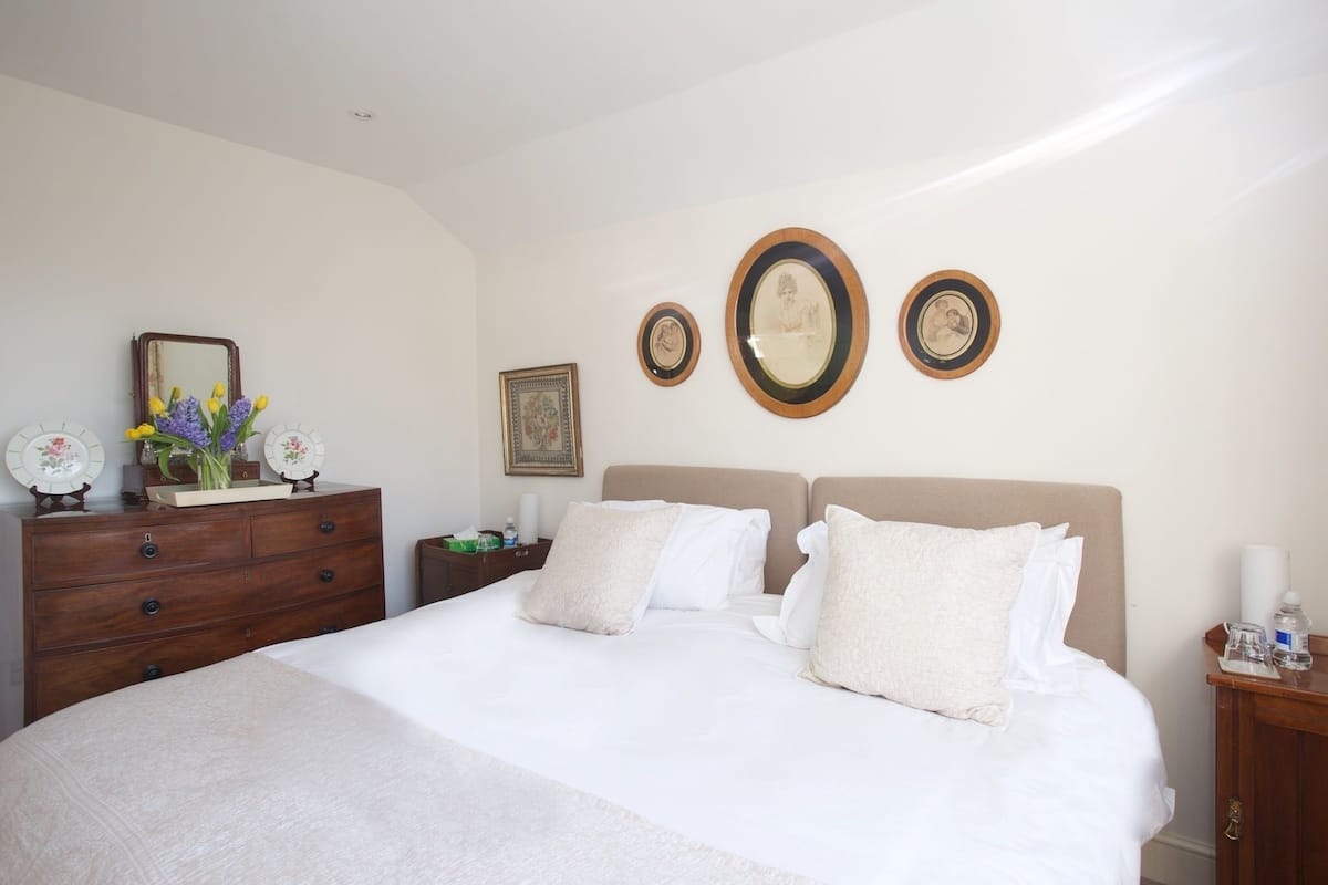Un dormitorio ordenado con una cama grande, un aparador y una mesita de noche. Hay tres cuadros en la pared, un espejo sobre el aparador y un florero con flores encima.