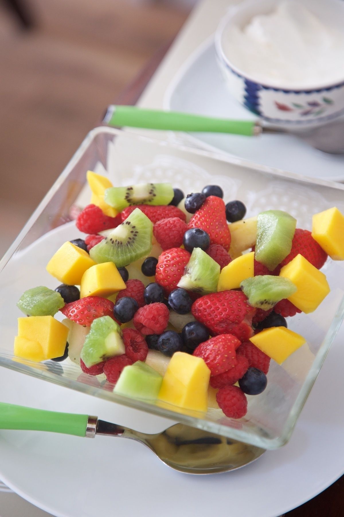 Un tazón de vidrio transparente lleno de frutas frescas variadas, como fresas, arándanos, frambuesas, kiwi y trozos de mango.