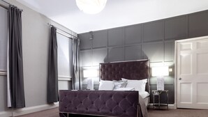 A bedroom with a large bed, a bedside table, a nightstand, a door, and a window with curtains. - The City Rooms (Leicester)