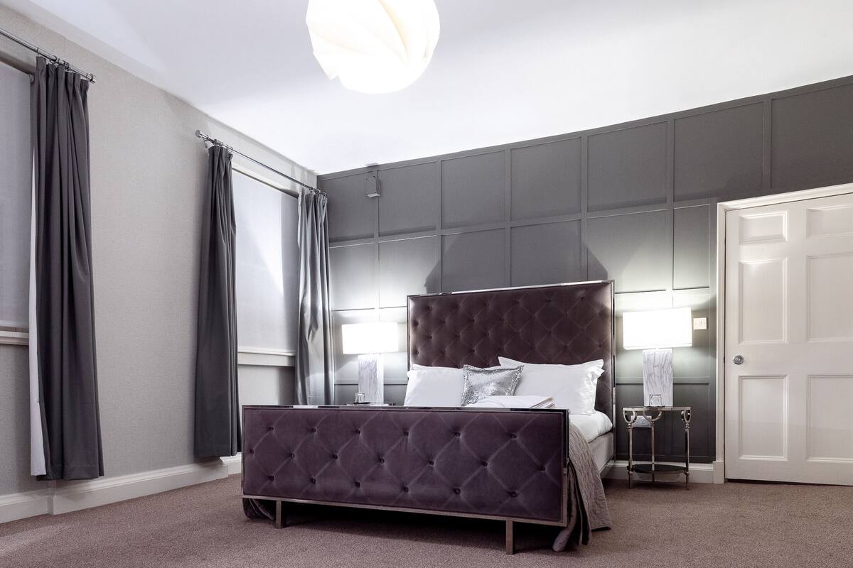 A bedroom with a large bed, a bedside table, a nightstand, a door, and a window with curtains.