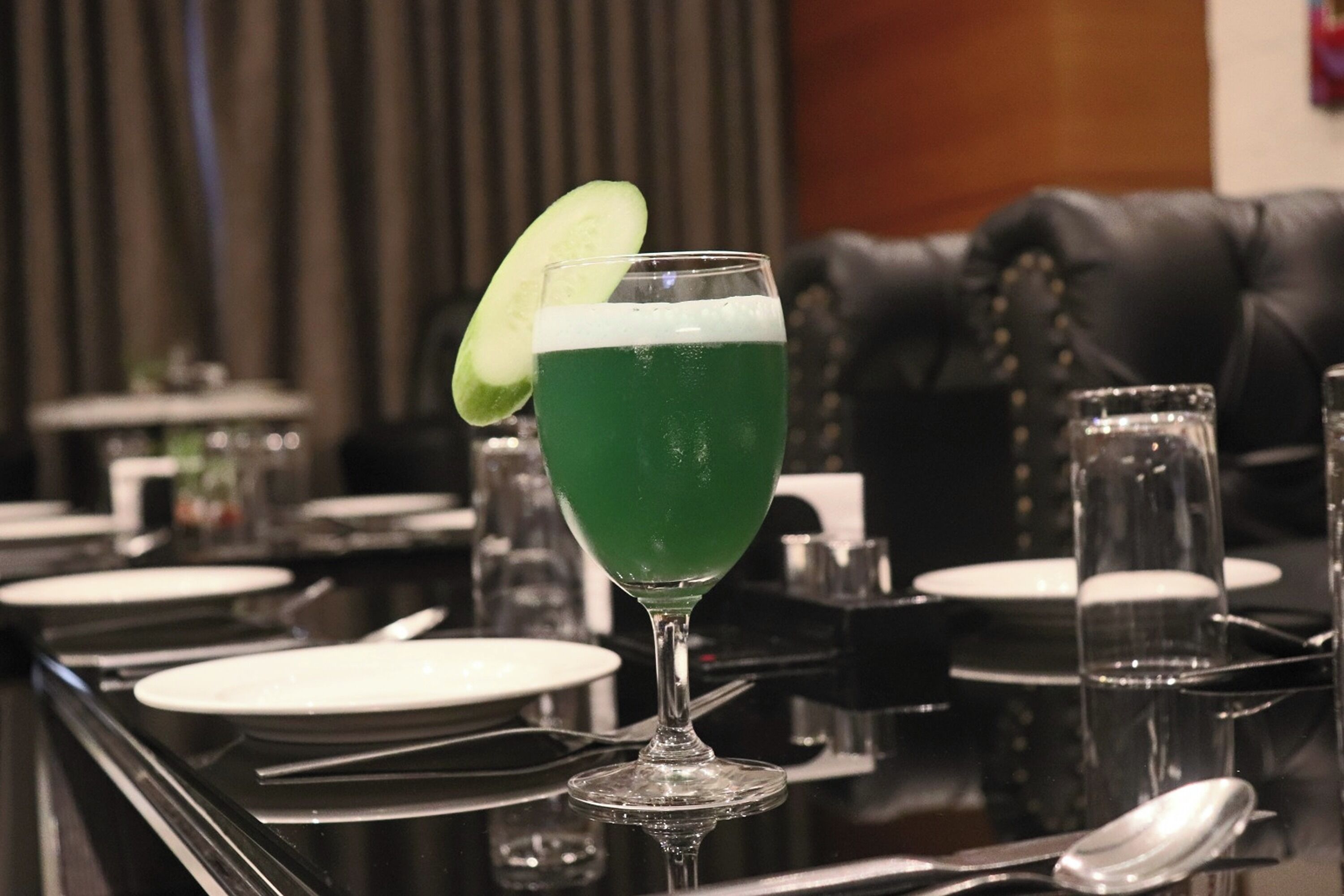 A bar setting with a green cocktail in a glass, a cucumber garnish, and a black leather couch in the background.