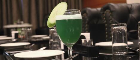A bar setting with a green cocktail in a glass, a cucumber garnish, and a black leather couch in the background.