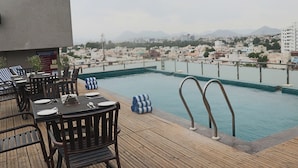 Zona de piscina en la azotea con terraza de madera y sillones de descanso, con vistas a un paisaje urbano que incluye edificios y montañas al fondo.