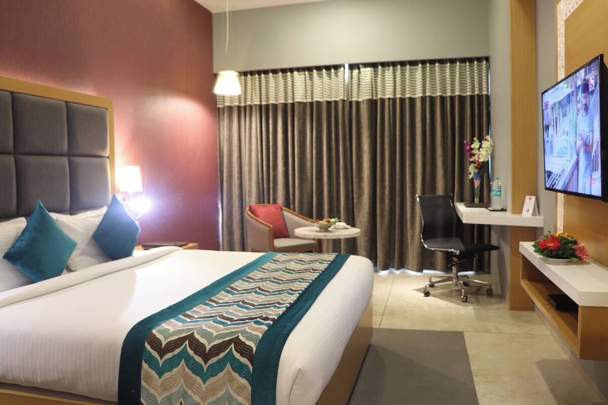 Una habitación de hotel con cama, escritorio, silla, televisor y ventana con cortinas.