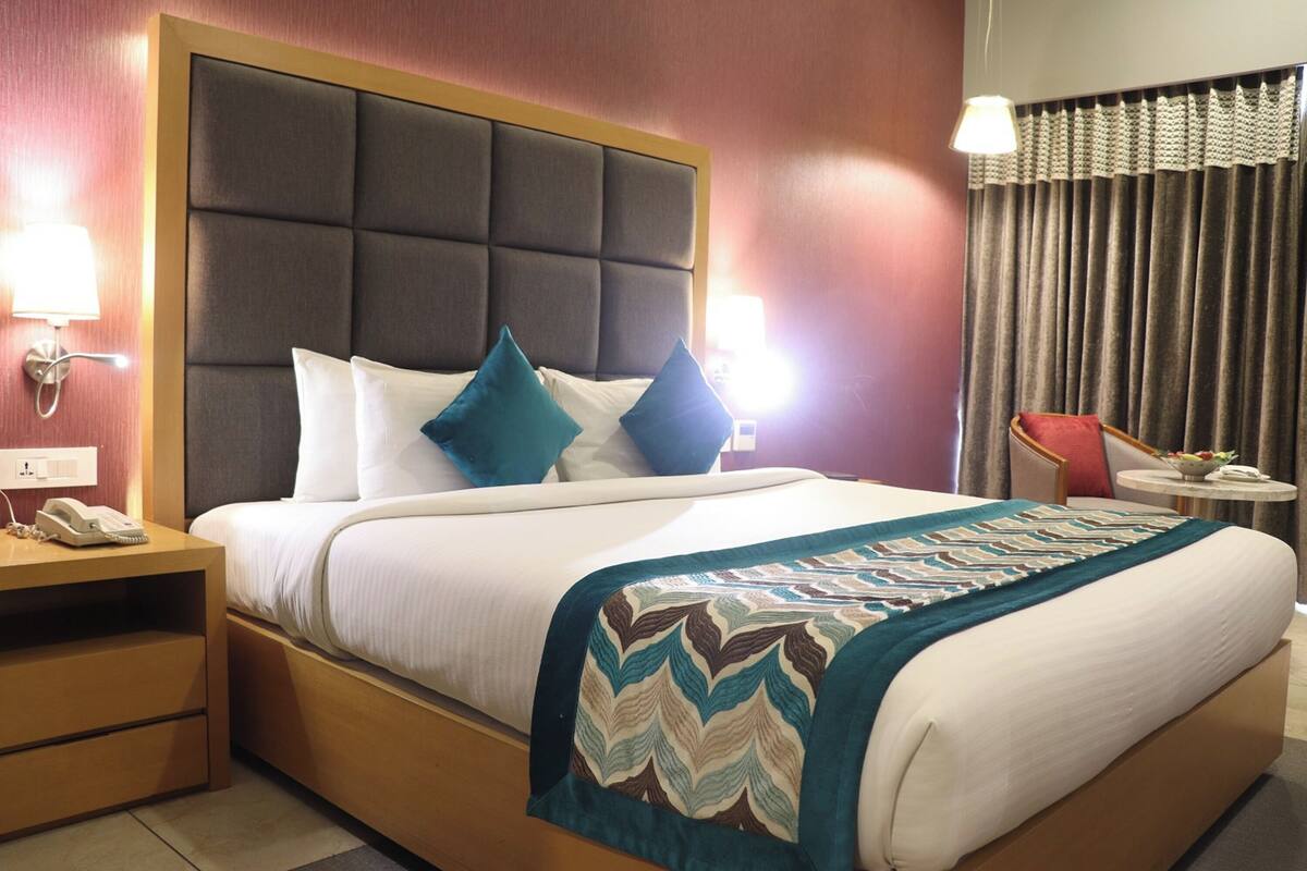 Una habitación de hotel con una cama grande, un cabecero, lámparas de noche, una mesita de noche y una pequeña mesa con una lámpara.
