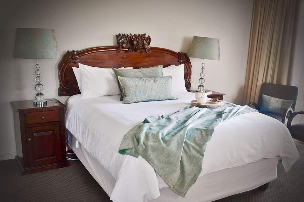 A neatly made bed with white linens and a green throw blanket, flanked by two bedside tables with lamps, a wooden headboard, and a chair in the corner.