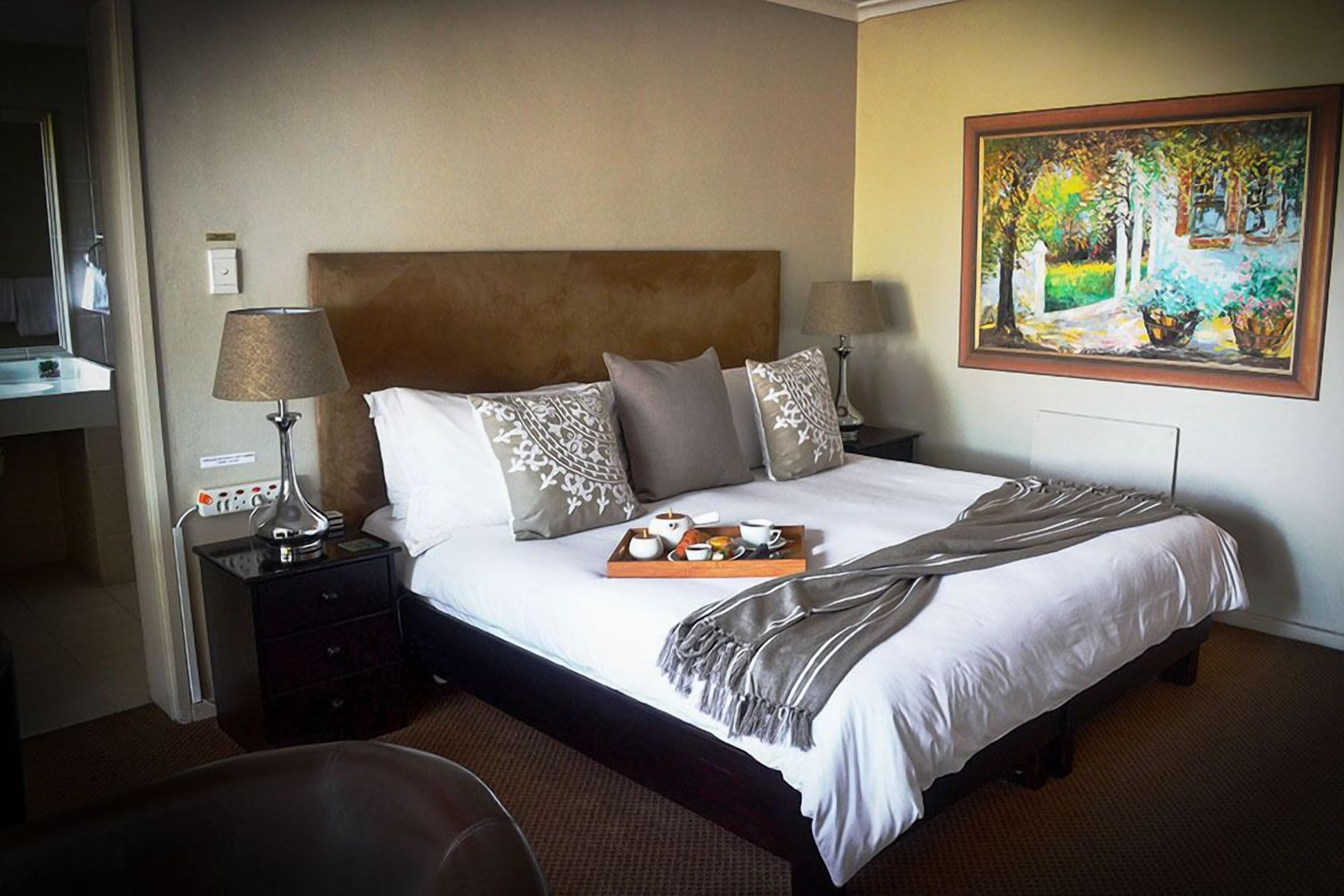 A hotel room with a large bed, two bedside tables with lamps, a painting on the wall, and a tray with breakfast items on the bed.