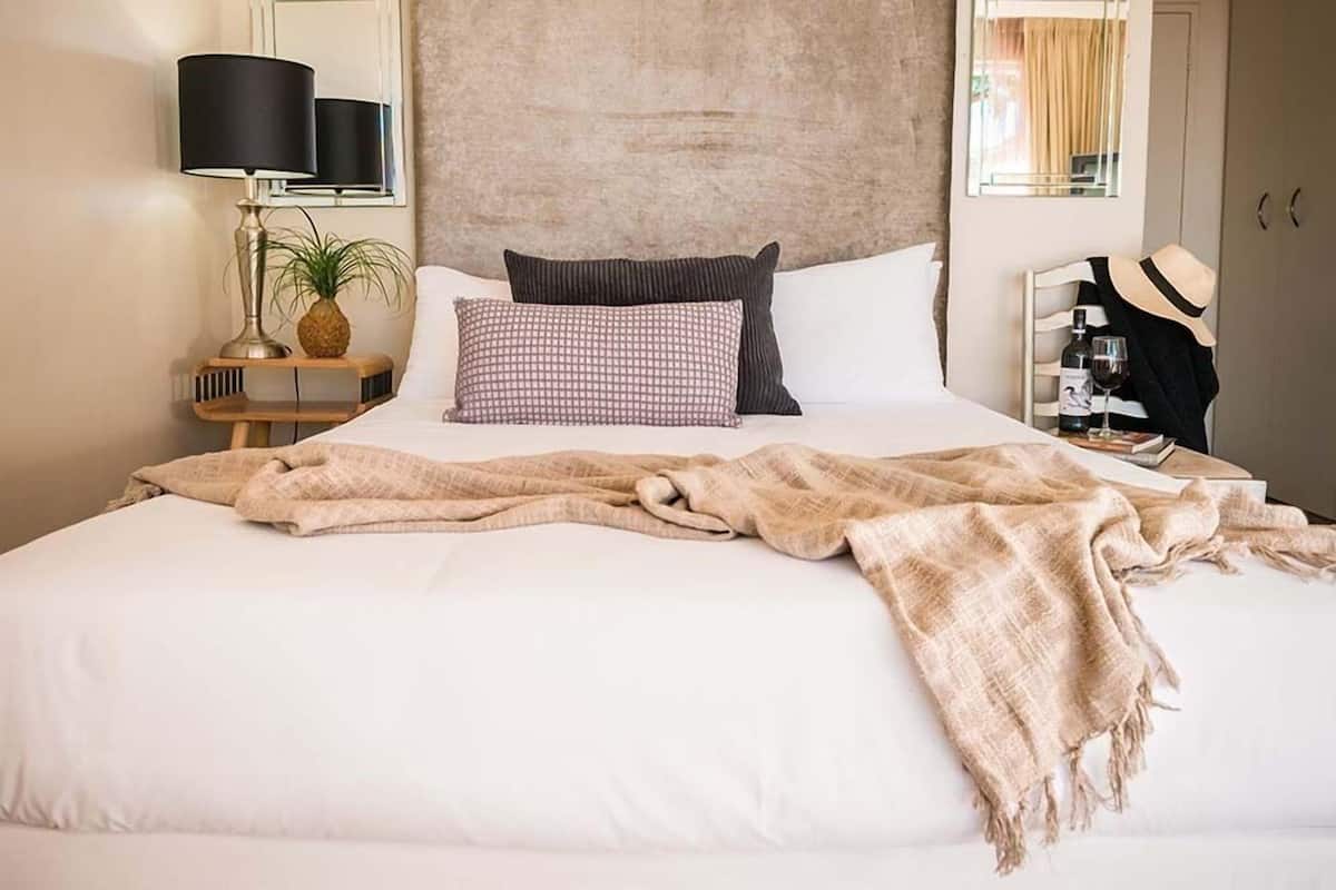 A neatly made bed with white linens, a beige throw blanket, and two pillows. There is a small wooden nightstand with a lamp and a plant beside the bed. A mirror hangs on the wall above the bed, and a hat and wine bottle are visible on a chair to the right.