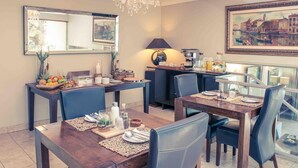 Un comedor con una mesa de madera preparada para cuatro personas, una cafetera y un frutero sobre una mesita auxiliar.