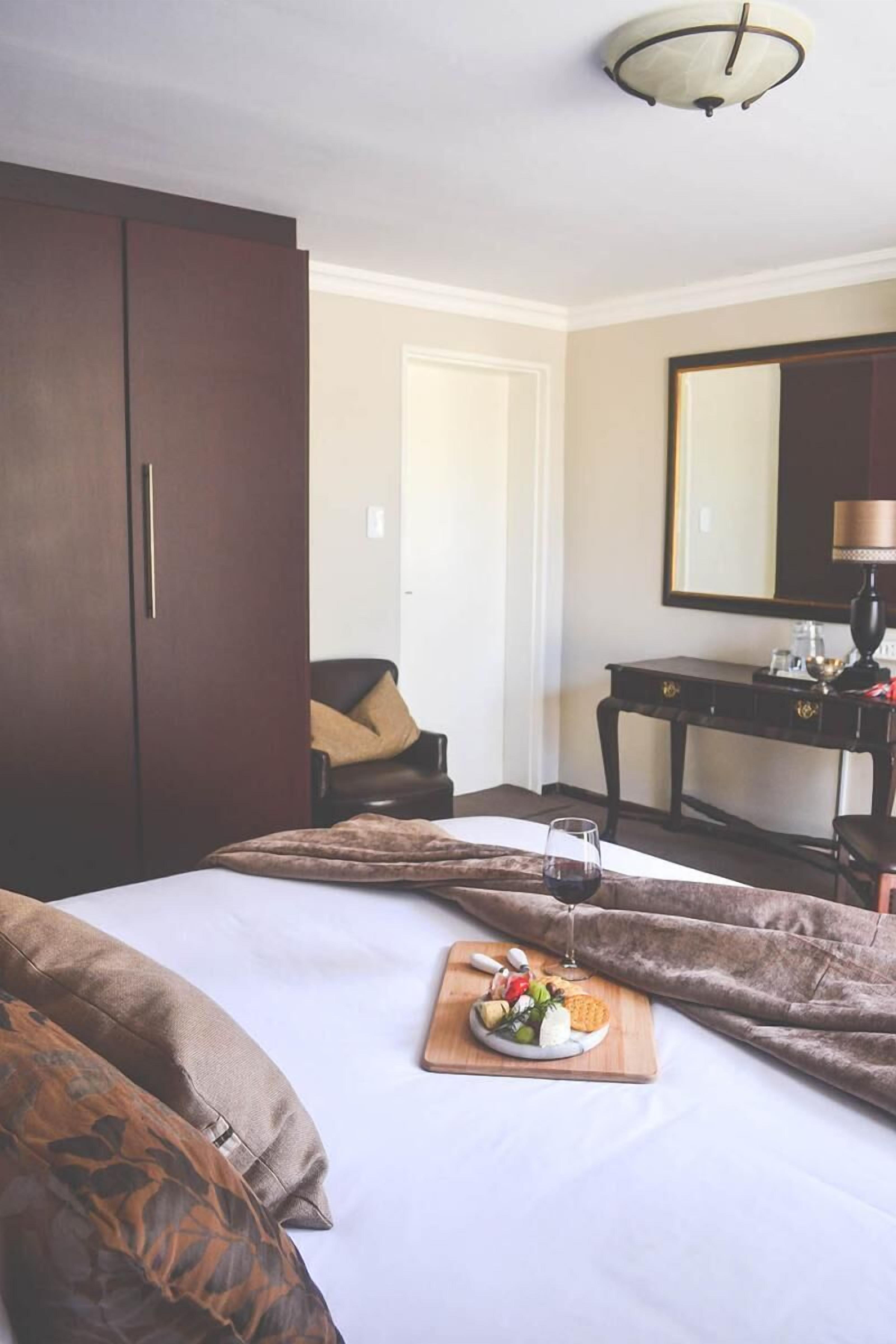 A hotel room with a bed, a bedside table with a lamp, a wardrobe, and a dining area with a plate of food and a glass of wine.