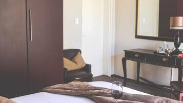 A hotel room with a bed, a bedside table with a lamp, a wardrobe, and a dining area with a plate of food and a glass of wine.