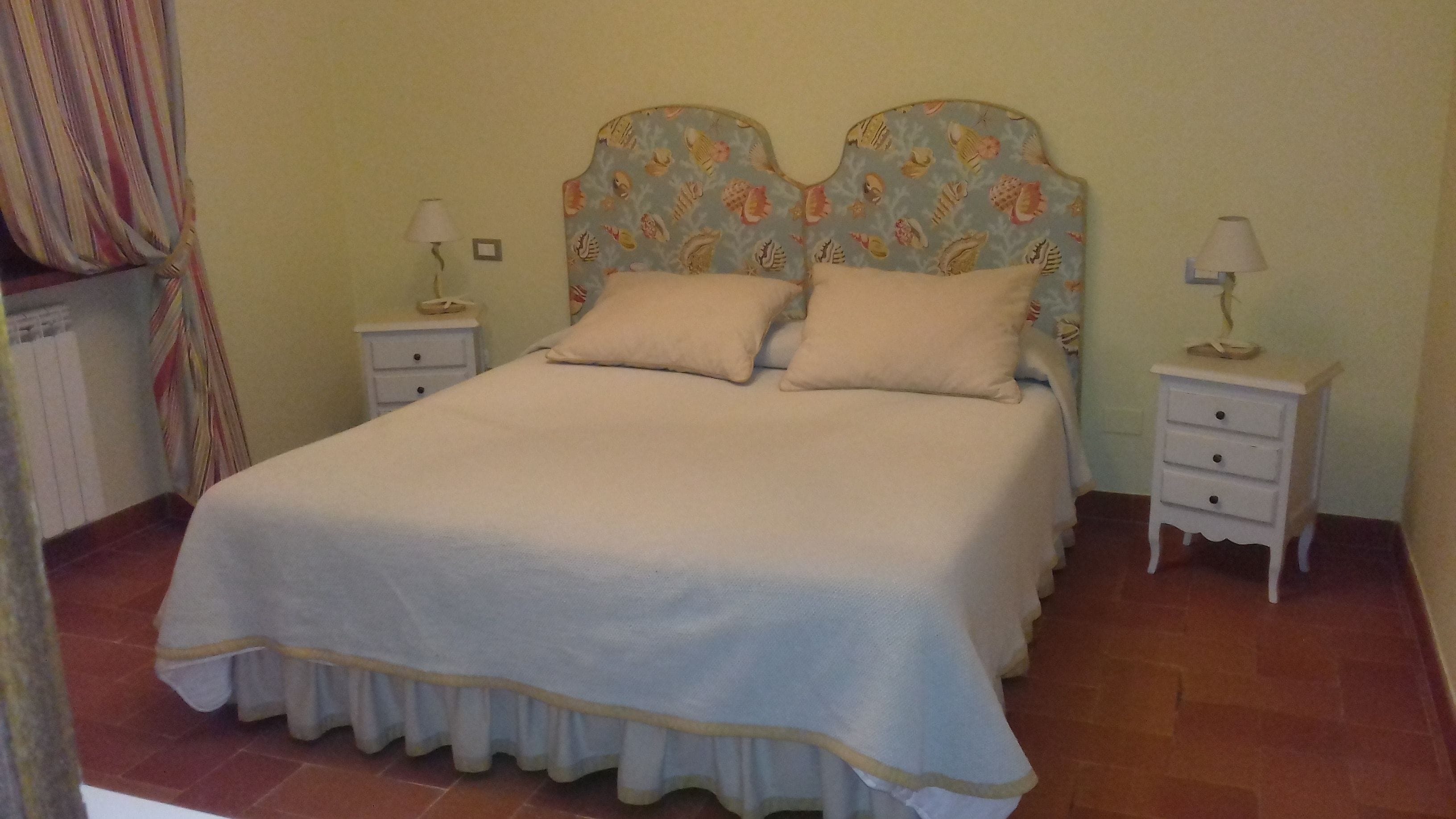 Un lit bien fait, avec une tête de lit à motifs, deux tables de chevet avec des lampes et un rideau sur le côté gauche.