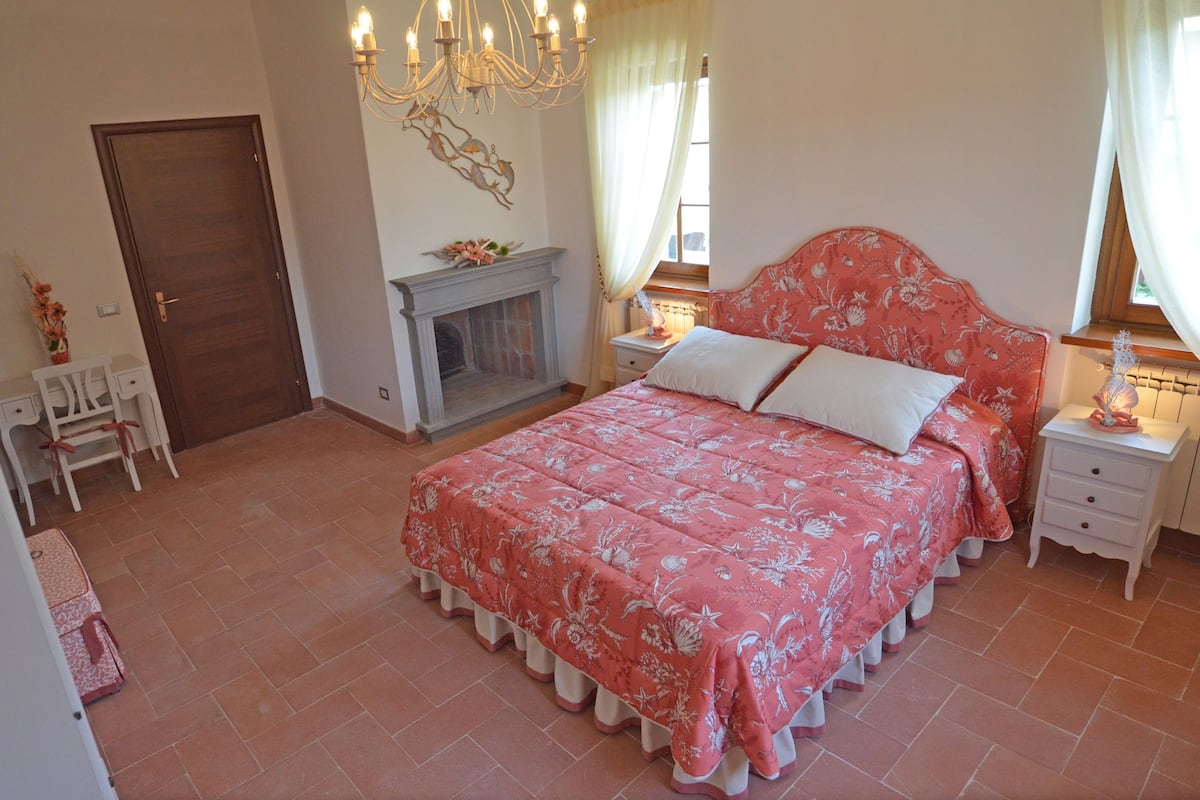 A bedroom with a large bed, a fireplace, a chandelier, and a nightstand with a lamp.
