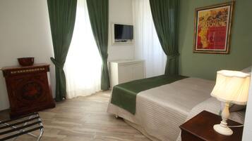 A bedroom with a bed, a nightstand with a lamp, a dresser, a television on the wall, and green curtains.