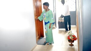 A woman in a patterned dress standing in a hallway with a wooden door, a man in a white shirt and jeans, and a flower arrangement on the floor. - Residence Rom 4 Treicheville (Abidjan)