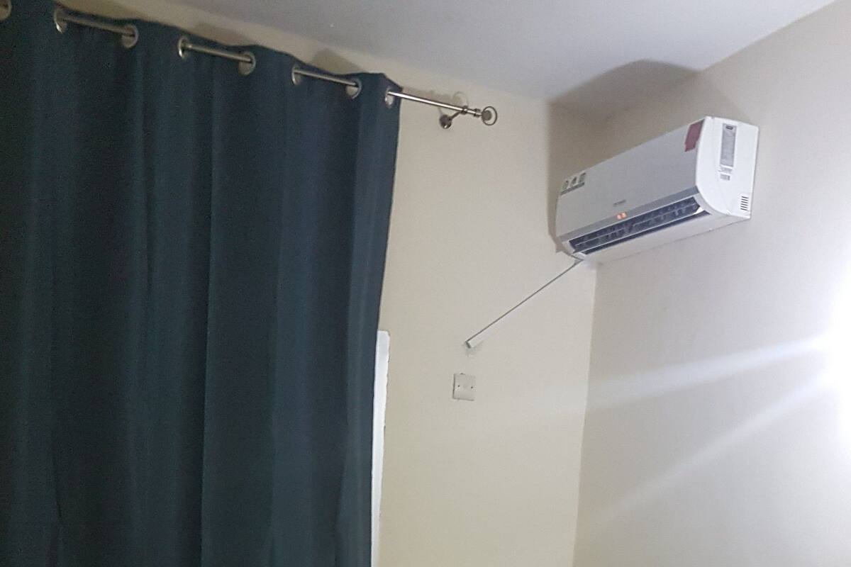 En säng med plats för en person, med vita sängkläder, en träfärgad sänggavel, en blå gardin och en väggmonterad luftkonditioneringsanläggning.