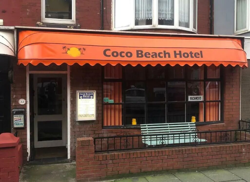Coco Beach Hotel with a bright orange awning, a visible window with a 'Vacancies' sign, and a bench outside.