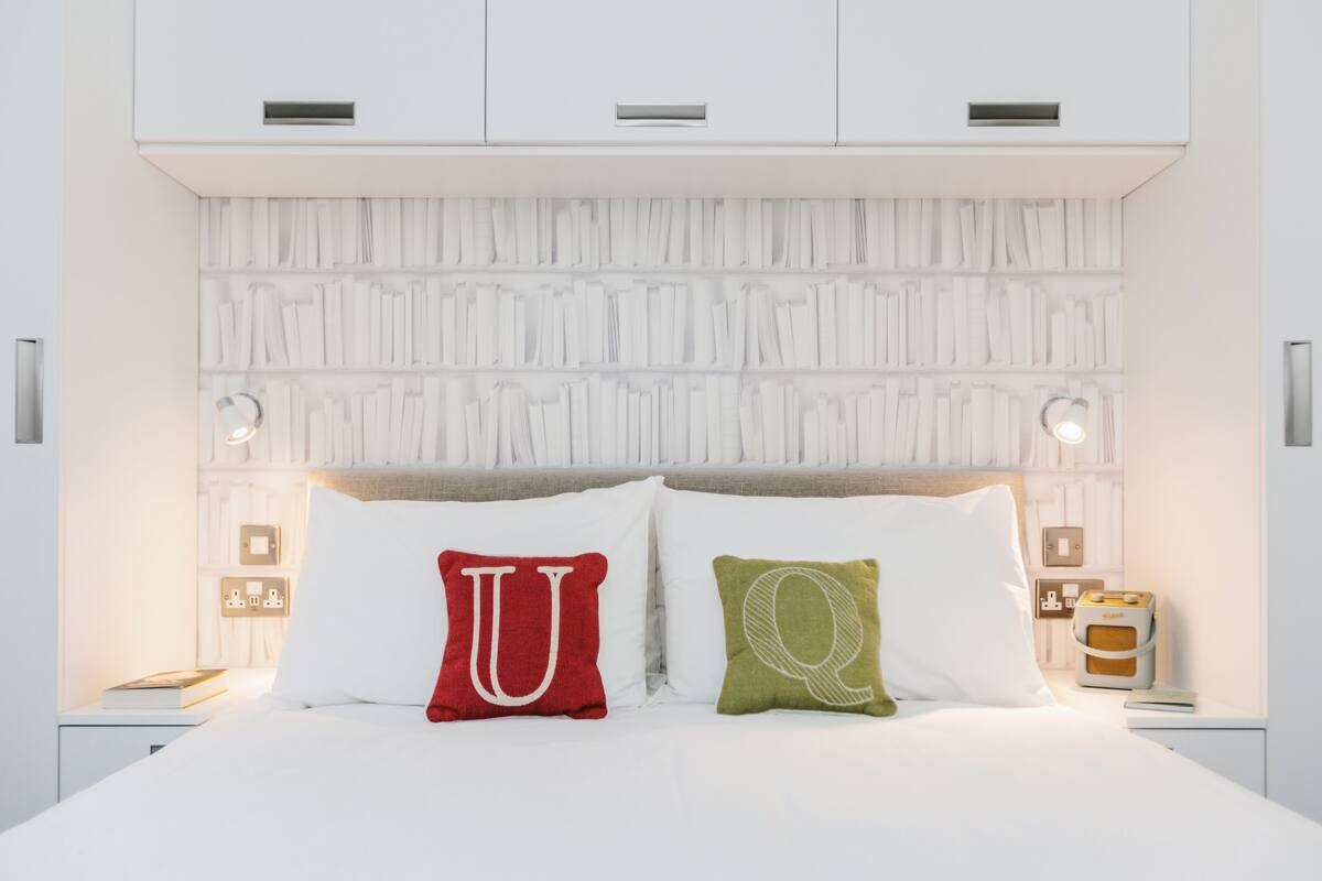 A modern bedroom with a white headboard, two bedside lamps, and a nightstand with a book and a small table.