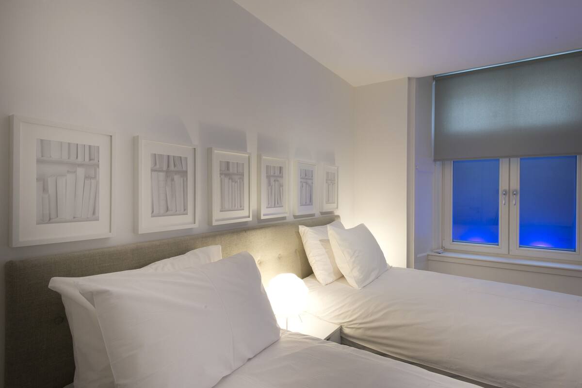 A hotel room with a bed, a headboard, a window with blinds, and framed artwork on the wall.